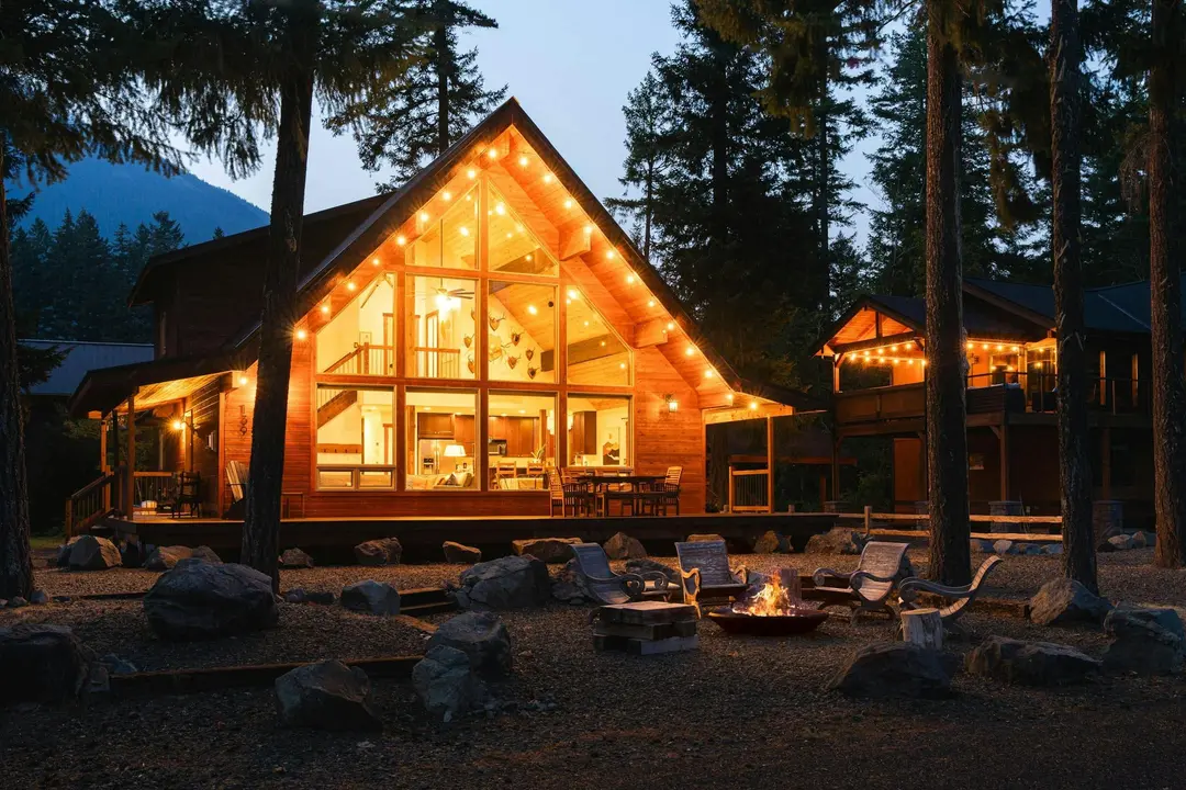 Holzhaus mit hohem, beleuchtetem Glasgiebel und Feuerstelle zur blauen Stunde zwischen Tannen